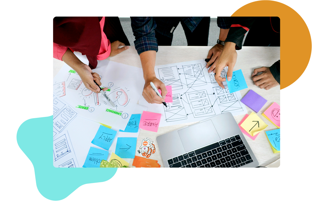 Overview of three people working on a table top with a laptop, paper, and sticky notes with strategy work on them.